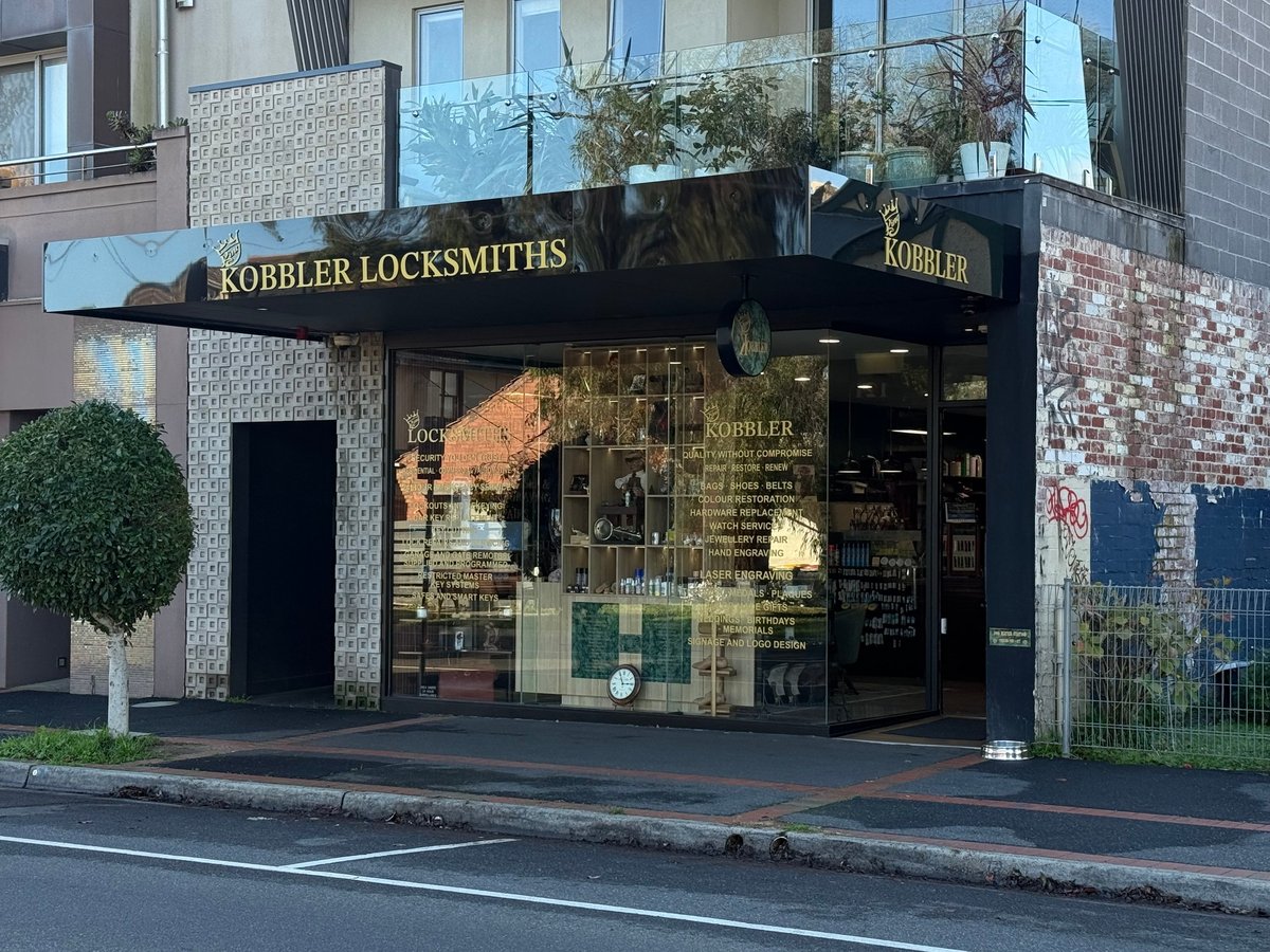 Exterior of Kobbler Locksmiths storefront with black awning, glass windows, and brick facade on a street corner